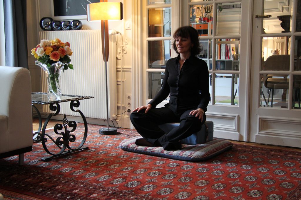 Une femme médite sur un coussin, dans le salon de son appartement parisien. Le décor est chaleureux.