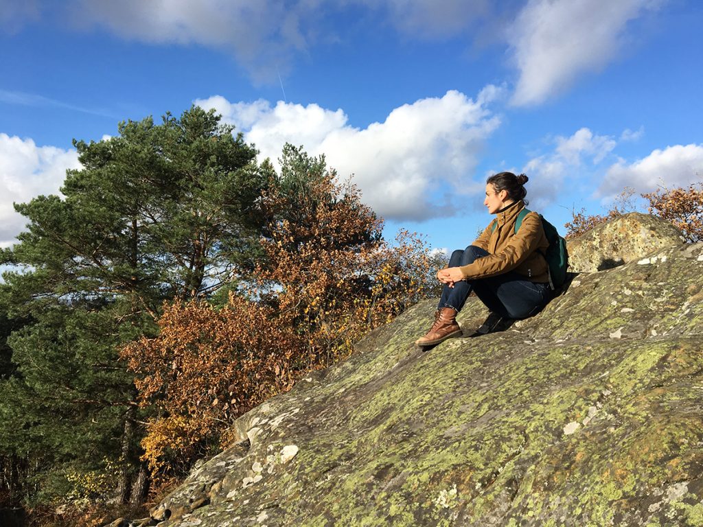 une jeune femme est assise sur un rocher en forêt de Fontainebleau. Elle regarde l'horizon. Derrière la nature et le ciel sont lumineux.