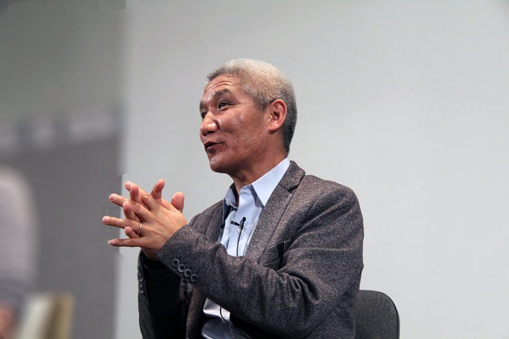 Portrait de Thupten Jinpa entrain d'ensigner. Il a le sourure aux lèvres et le smains jointes devant lui pour montre un sens d'unité.