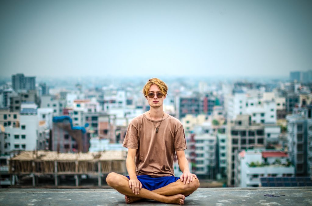 Un jeune homme est assis en méditation au coeur d'une capitale urbaine. Il est en short et porte des lunettes de soleil.