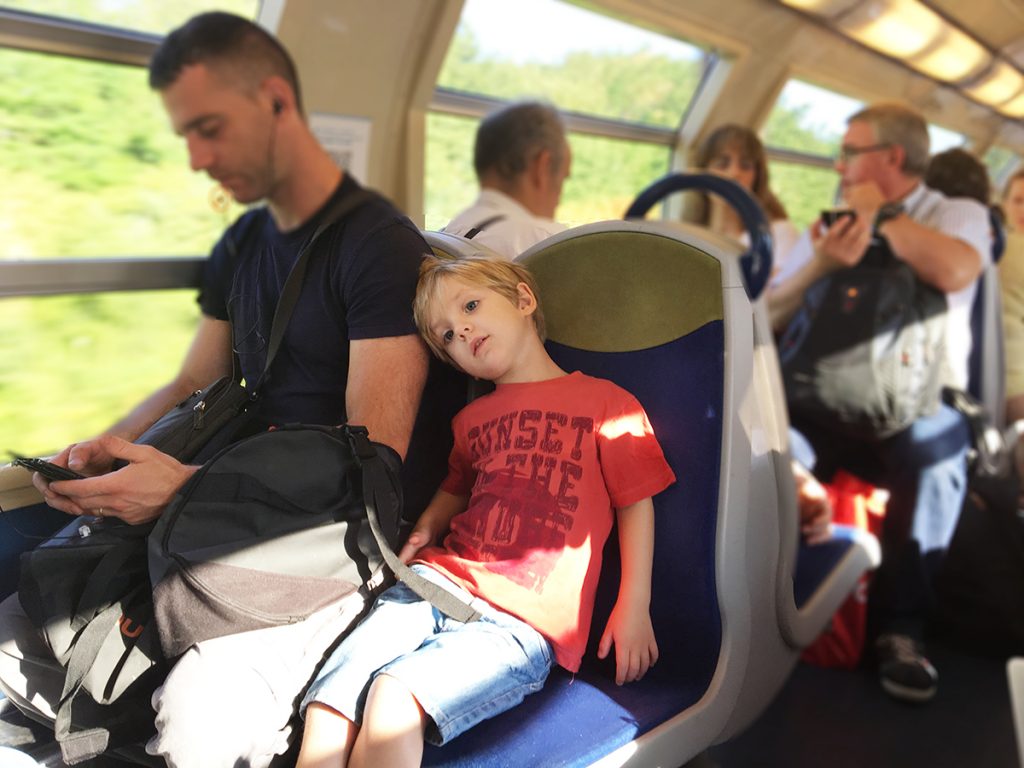 Un enfent dans le RER francilien, porte un tee-shirt. Il est appuyé contre son père qui regarde son téléphone. Tout le monde à chaud.