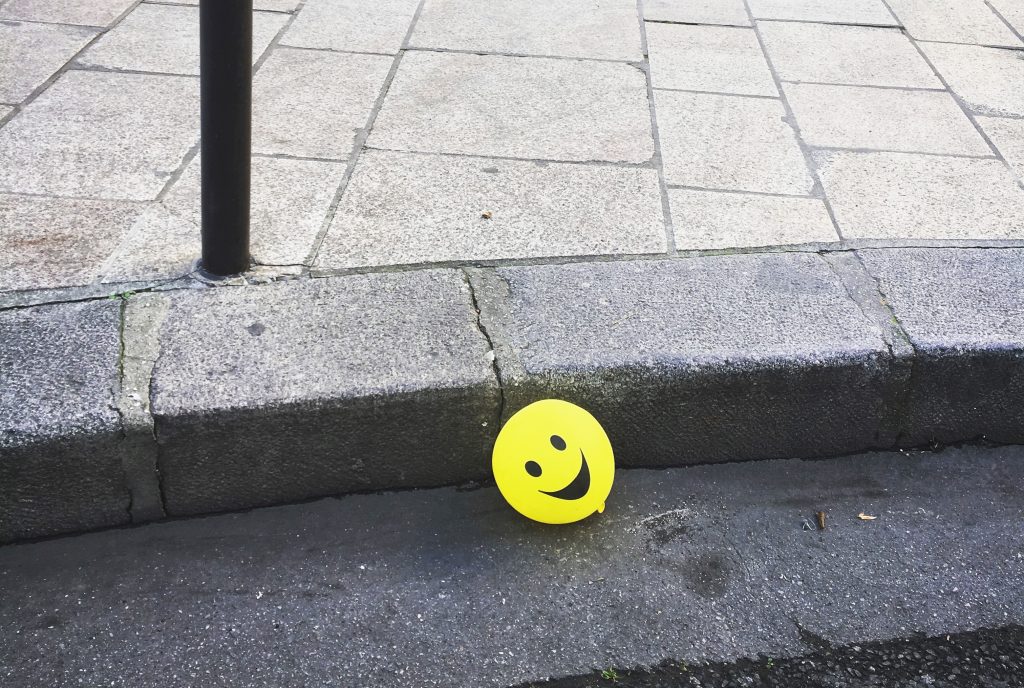 Un ballon jaune avec smiley est tombé par terre sur la chaussée grise. Ce sourire vif contraste avec la couleur du bitume. Un moment de tendresse