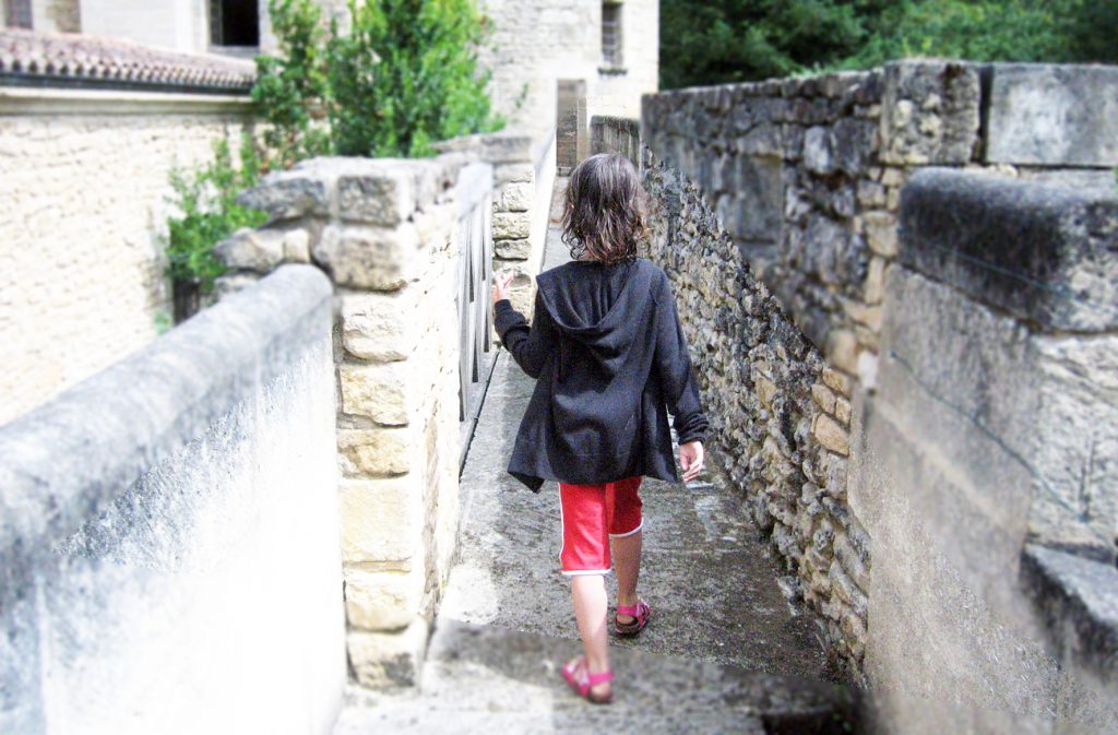 une petite fille marche sur un chemin de pierre. on la voir de dos. Elle porte un sweat noir et un pantacourt rouge.