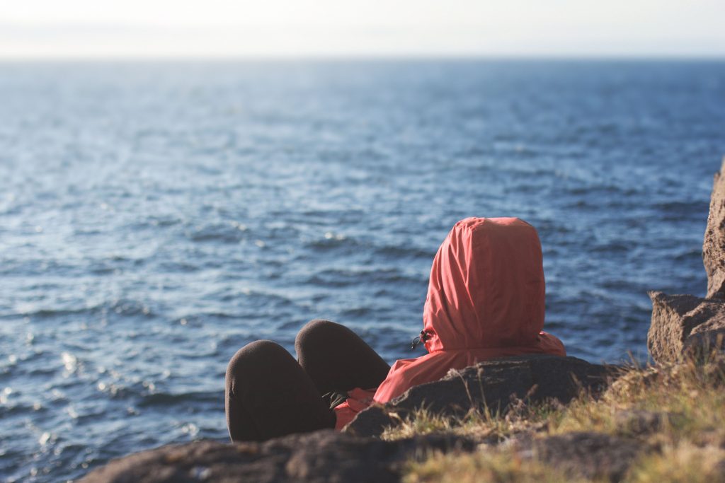 un homme (ou une femme) vêtu d'un Kway rouge, assis face à la mer