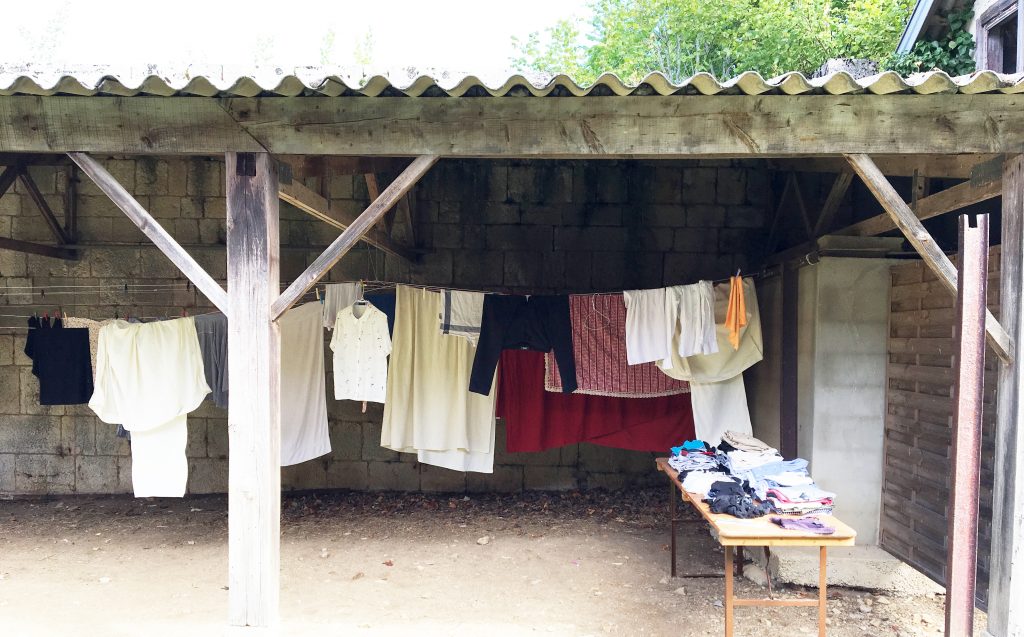 Sous un auvent, une lessive est étendue. Du linge plié est posé à droite des fils à linge, sur une table.