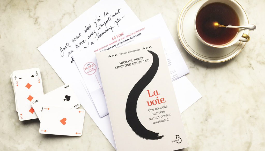 le livre La Voie est posé sur une table en marbre à côté d'une tasse de café et d'un jeu de cartes