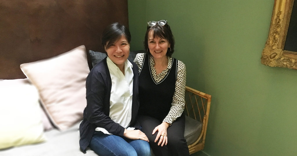 Regina Wong et Marie-Laurence Cattoire sont assises ensemble sur un canampé en rotin, dans le lobby du petite hôtel parisien Le Coq Hotel. Elles sourient toutes deux à l'objectif. Elles sont vêtues assez simplement en noir et banc. Le mur derrière elles est d'une jolie couleur verte.