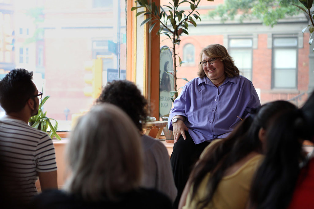 Sharon Salzberg donn une conférence. Elle sourit aux auditeurs. la salle est lumineuse, éclairée par une large baie vitrée.