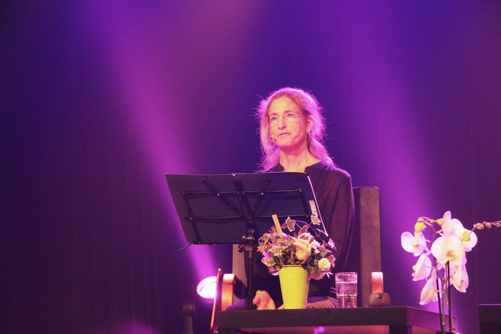 Tara Brach sur une scène de théâtre, baignée de lumière violette, donne une conférence à Paris. Devant elle un lutrin supporte le texte de sa conférence. Devant elle un bouquet de fleurs dans un vase jaune