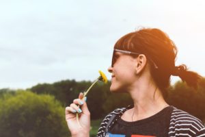 Nourrir nos cinq sens. Une femme souriante, de profil, avec une queue de cheval, regarde le ciel en sentant une fleur jaune