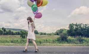 Une jeune femme en robe blanche marche sur une route de campagne. Elle tient des ballons gonflables de toutes les couleurs