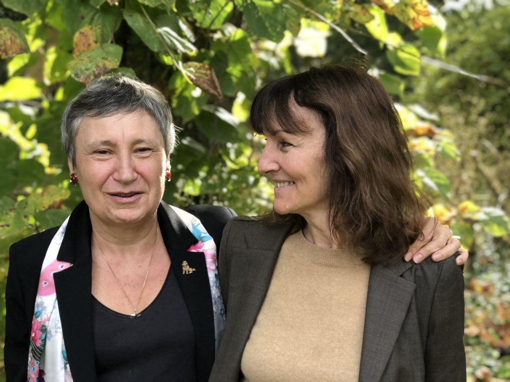 Marie-Laurence Cattoire regarde l'auteure Danielle Moyse en souriant. Elles se tiennent par les épaules et posent à l'extérieur, dans un jardin, devant des feuilles d'arbres ensoleillées.