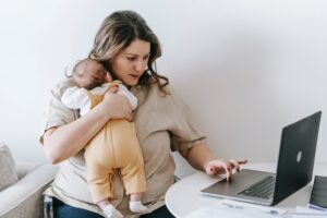 Une jeune maman porte son nourrisson sur l'épaule tout en essayant d'écrire sur son ordinateur portable