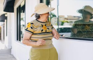 Le tabou de l'âge des femmes. Femme âgée portant un chapeau et riant de voir son reflet dans une vitrine. Elle est habillée de couleurs vives et porte un rouge à lèvres rouge vif