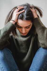 Une jeune femme se prend la tête dans les mains. Elle semble être assaillie par le doute. Elle porte un pull vert et un jean