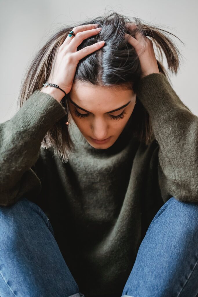 Une jeune femme se prend la tête dans les mains. Elle semble être assaillie par le doute. Elle porte un pull vert et un jean