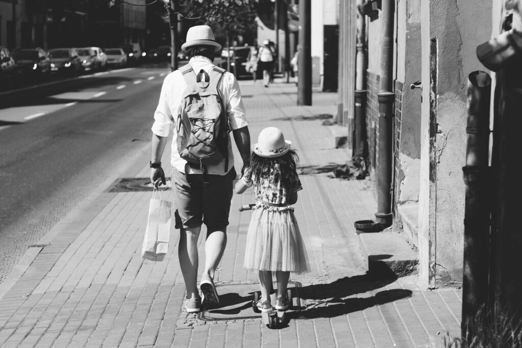 UN père tient la mains de sa petit fille.iLs marchent dans la rue sous un beau soleil. La photo est en N&B, on les voit de dos.