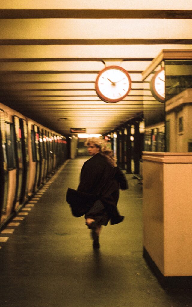 Une jeune femme en robe et manteau noirs court sur un quai de métro. Elle tourne le regard vers l'objectif prête à changer d'avis. La photo est en couleur, prise le soir. Un egrosse pendule indique l'heure, accrochée au plafond.